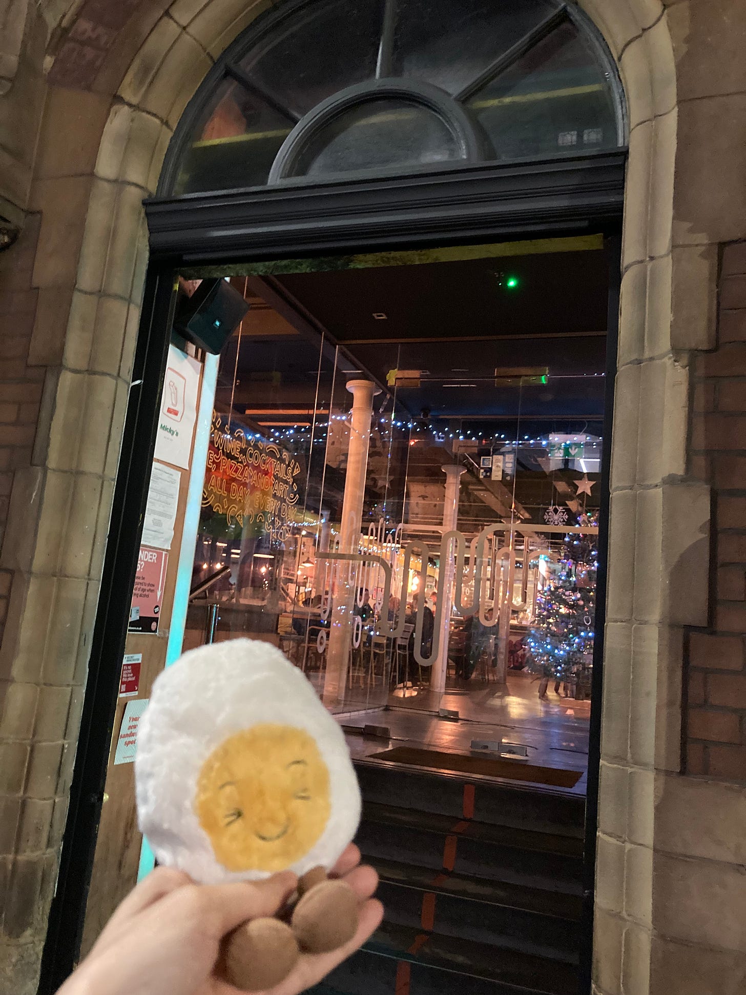 Photograph of dippy the soft toy egg outside of a pizza place with a stonework arch glass doorway showing a christmas tree and lights inside. 