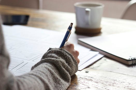 erson writing with a pen on paper, at a desk, with a coffee cup at the end of the desk.