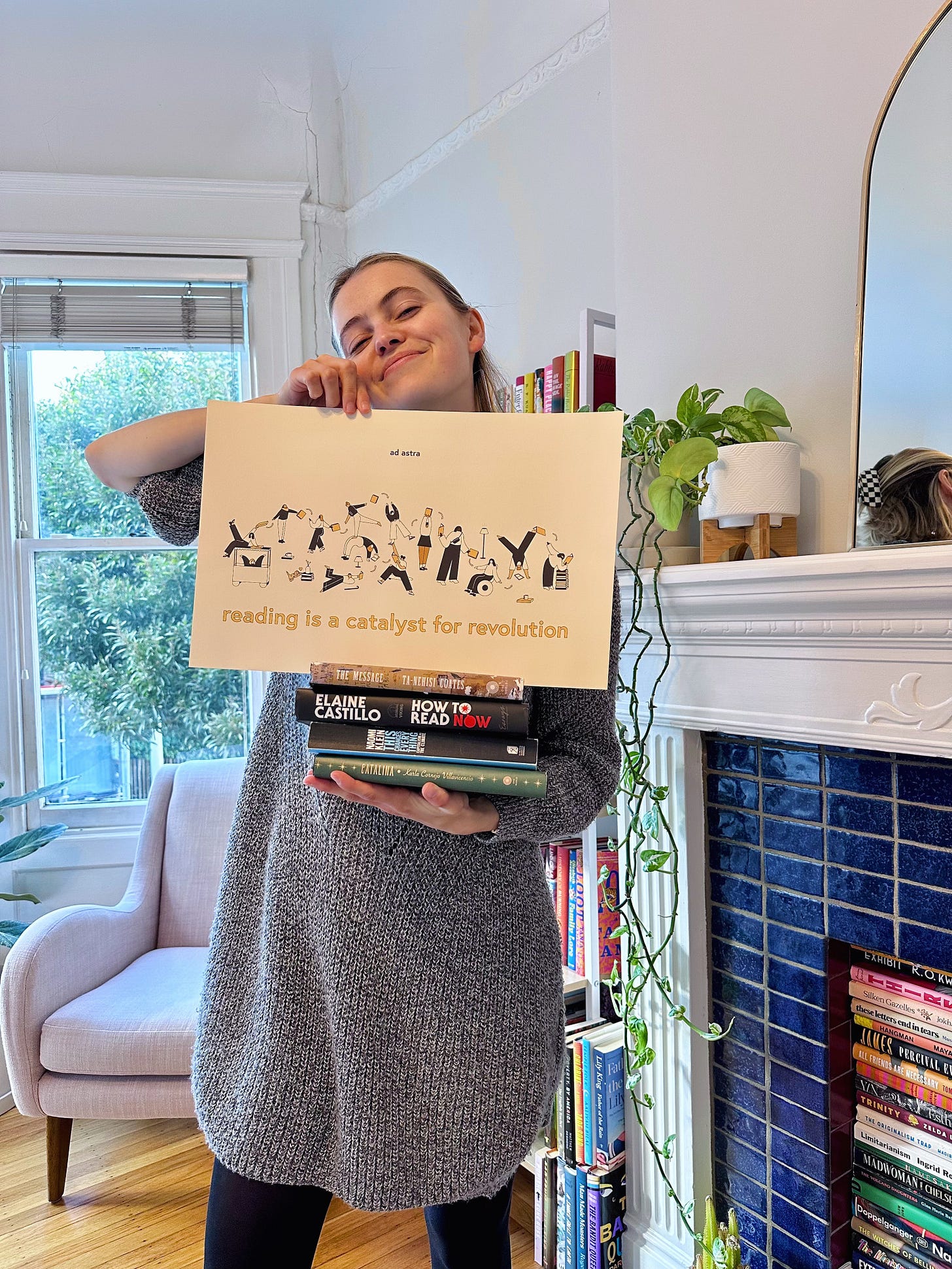 A blonde woman holds a stack of books with a print resting on top that reads: reading is a catalyst for revolution