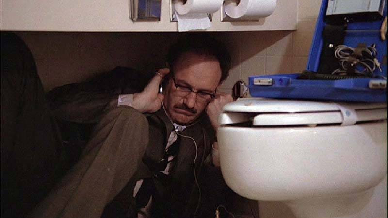 Screenshot from the movie The Convsersation showing Harry Caul listening on a surveillence device balanced on a toilet.