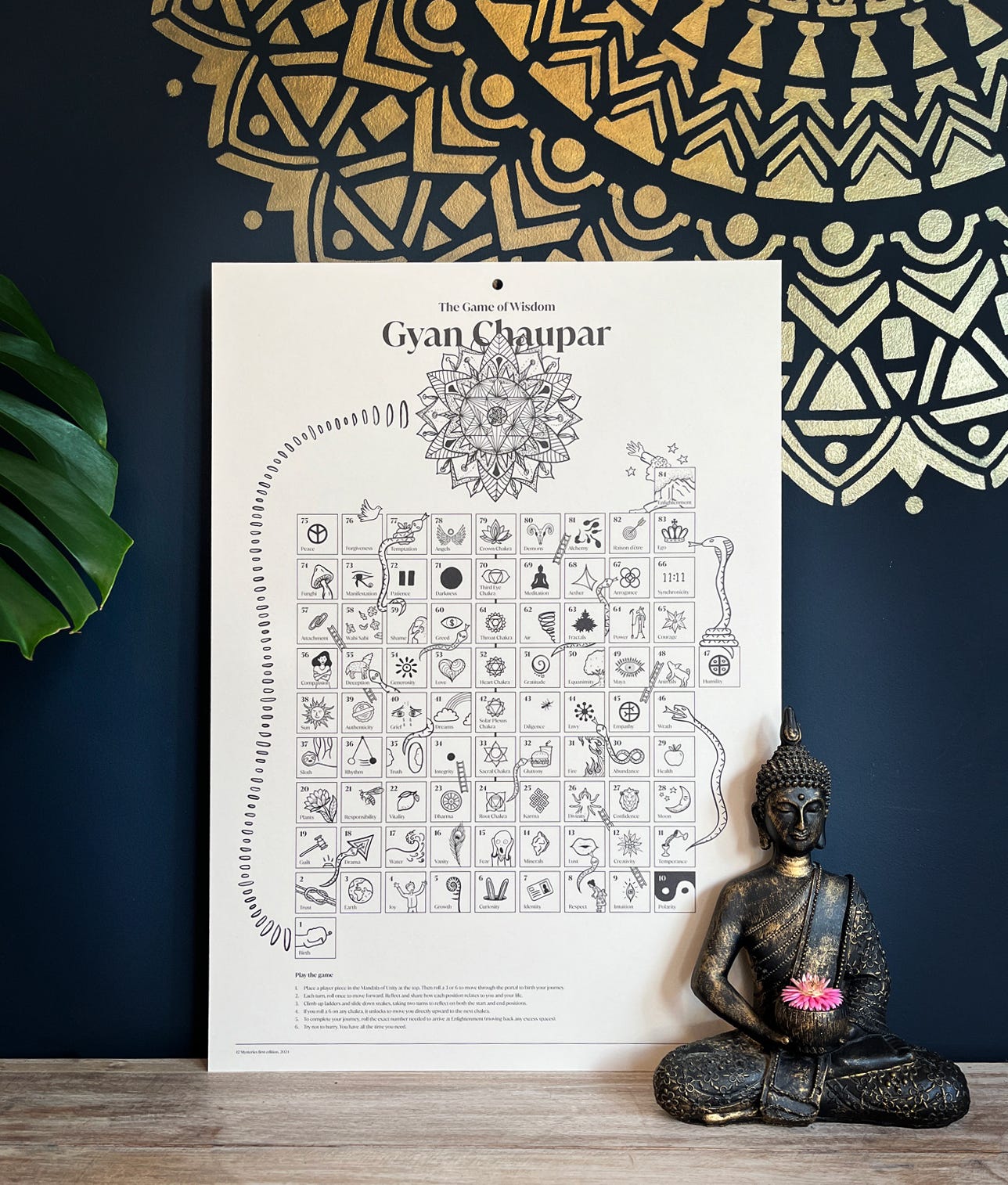 A Gyan Chaupar game board leaning against a dark blue wall with buddha statue in the foreground