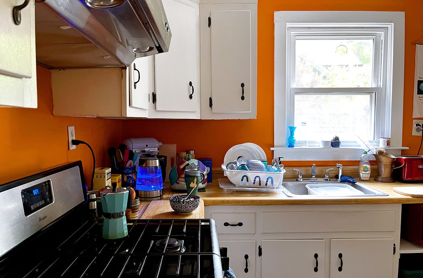 bright orange walls and ancient white kitchen cupboards, kitchen clutter includes turquoise glassware and coffee pot