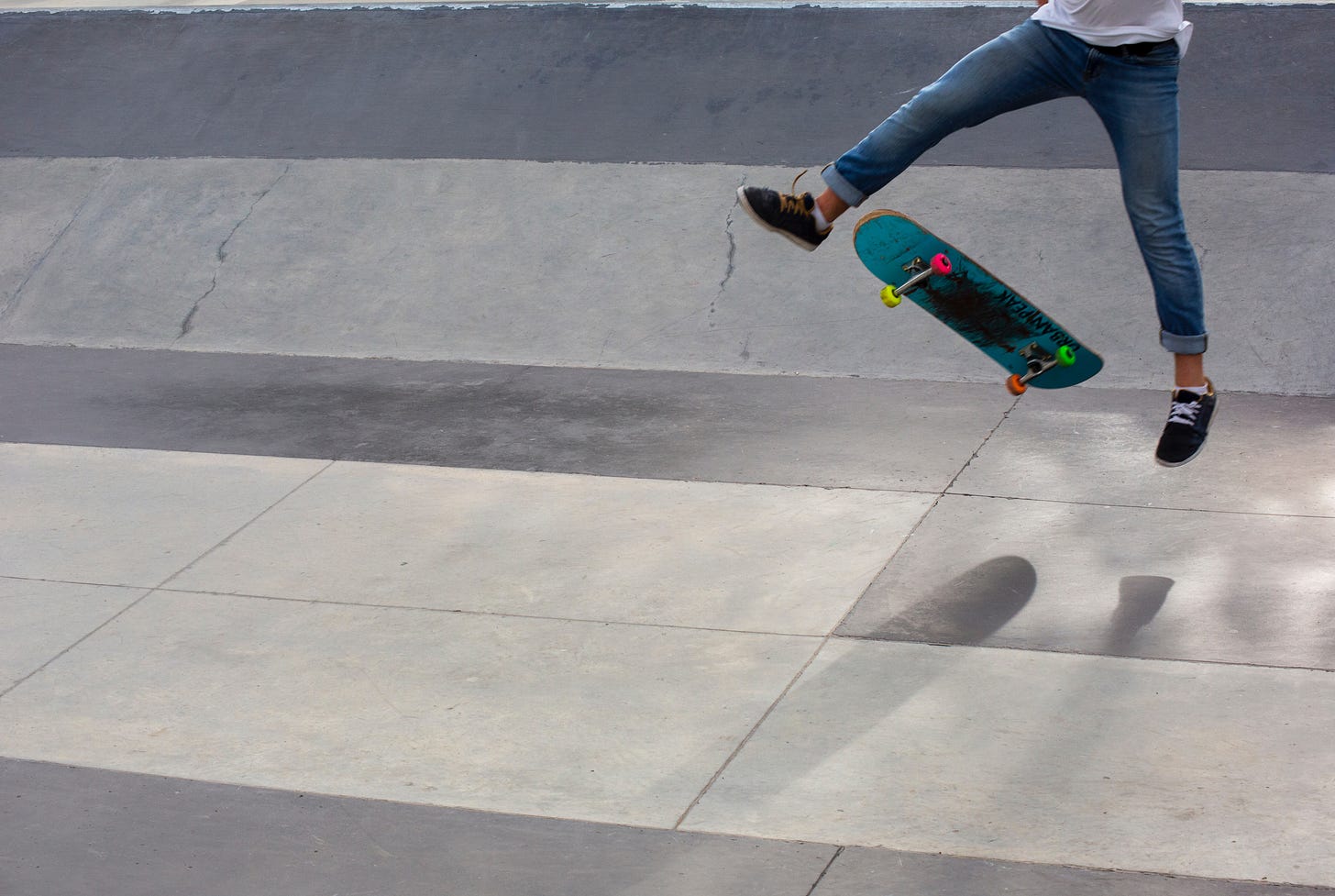 In the picture, the lower half of a person is seen in the upper right, jumping in the air. Between their legs is a blue and black skateboard. The person is wearing jeans and black shoes. The ground beneath them is gray and white concrete.