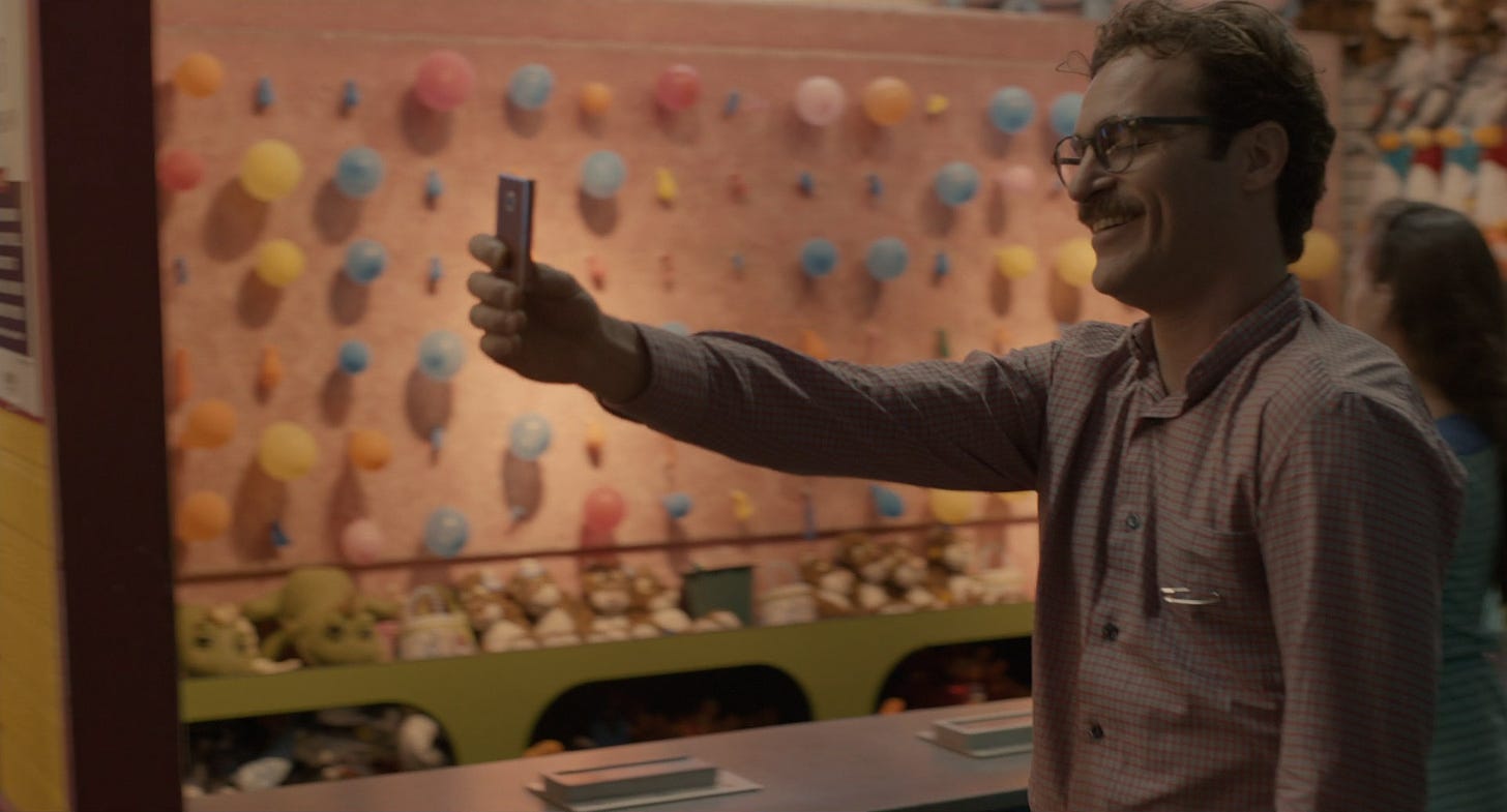 cena do filme Her. homem branco de óculos e camisa rosa segura celular na frente do rosto e sorri. No fundo, uma banquinha de parque de diversões com balões numa parede