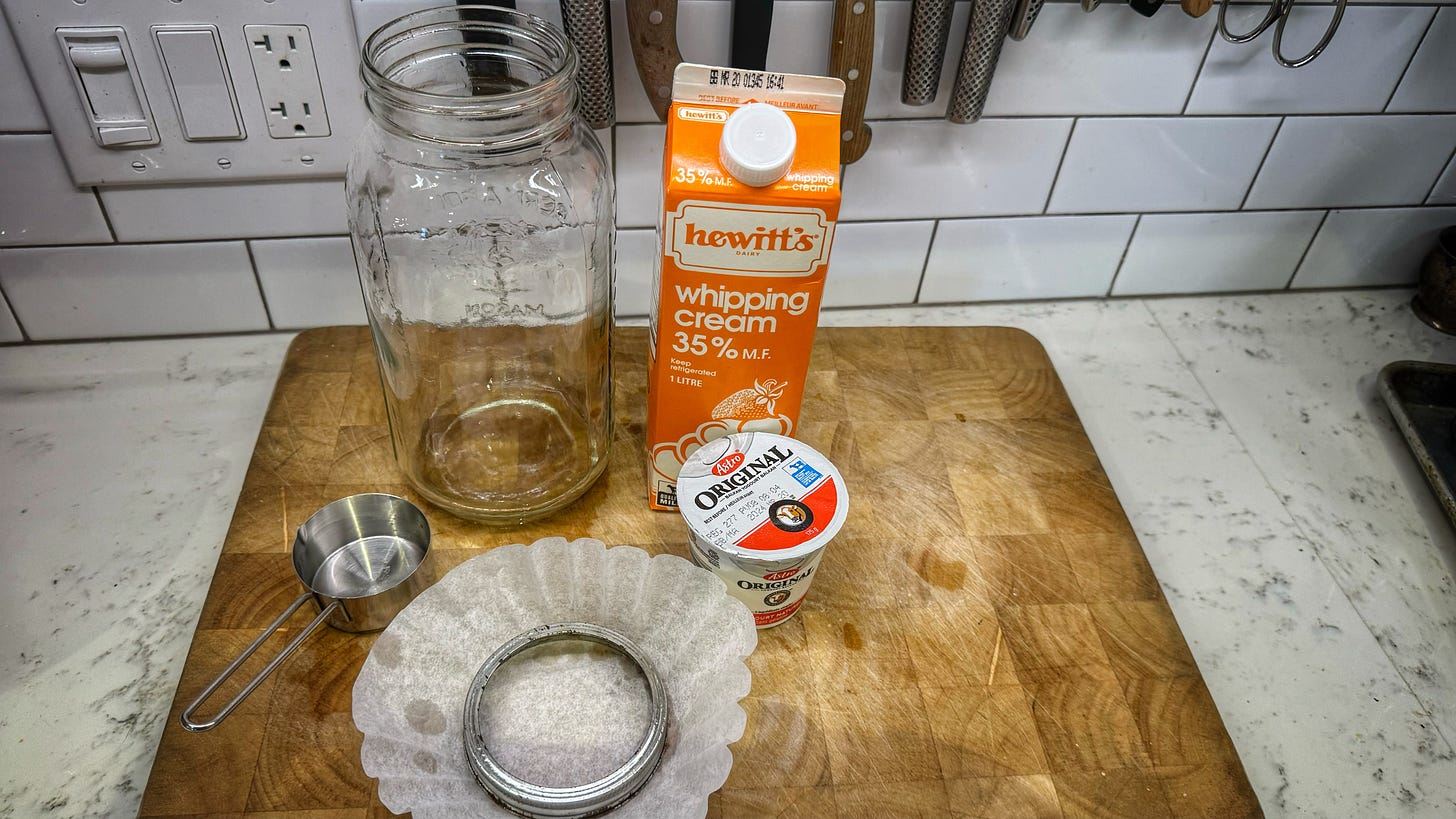 A very large mason jar, a litre of 35% whipping cream, a single-serving pot of yougurt, a coffee filter, the mason jar's twist band, and a 1/3 cup measuring cup, all on a cutting board. 