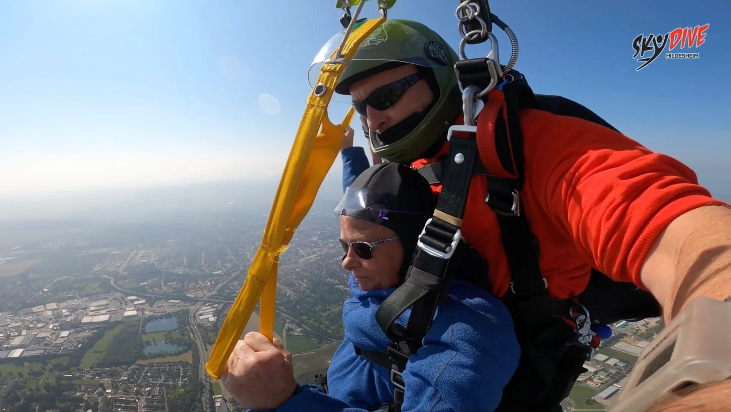 Tandem-Fallschirmspringen über Hildesheim, Deutschland: Eine Person wird von einem Instruktor in einem orangefarbenen Anzug und grünem Helm aus dem Flugzeug begleitet, während sie unter einem gelben Fallschirm schwebt. Die Aussicht auf die Stadt und die umliegende Landschaft sind klar zu erkennen, und die Person hält sich am Fallschirm fest, während sie sich dem Gefühl von Freiheit hingibt.