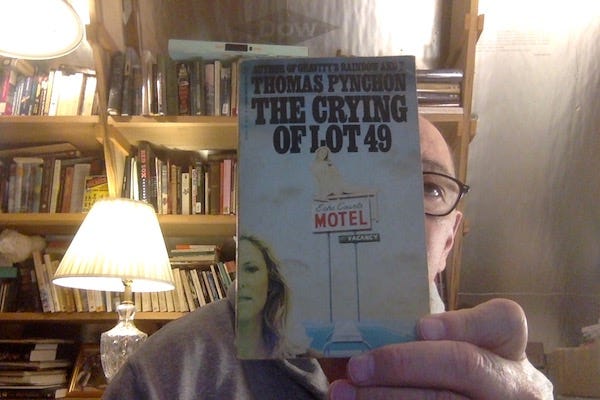 Photo of a some guy in a dimly lit, book lined office holing a copy of The Crying of Lot 49 to the camera. The book mostly obscures his face. 