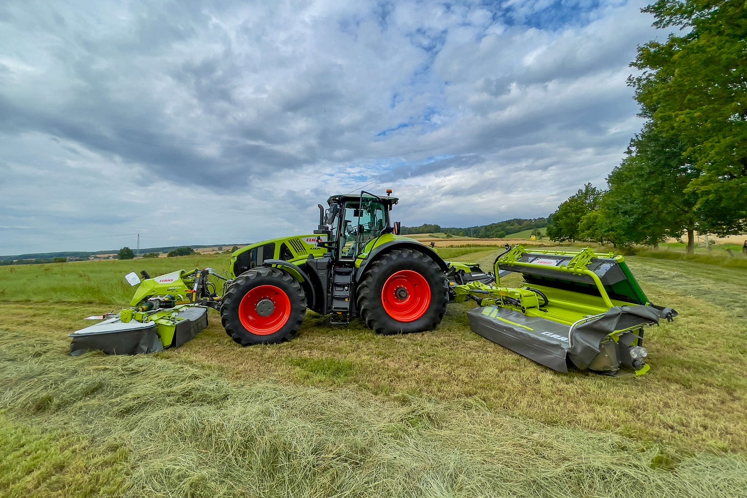 Ein grüner Claas-Traktor mit roten Rädern steht auf einer gemähten Wiese, ausgerüstet mit Mähwerken vorne und an den Seiten. Der Himmel ist bewölkt, im Hintergrund erstrecken sich Felder und Bäume. Ein grüner Claas-Traktor mit roten Rädern steht auf einer gemähten Wiese, ausgerüstet mit Mähwerken vorne und an den Seiten. Der Himmel ist bewölkt, im Hintergrund erstrecken sich Felder und Bäume.