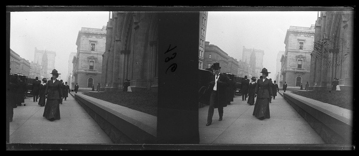 People in formal dress walking along sidewalk outside stone church or civic building, with women in long skirts and men wearing bowler hats and overcoats.