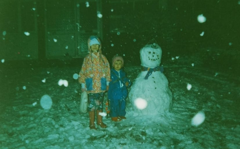 Two young girls in brightly coloured coats standing next to a big snowman at night time. It's snowing in the picture