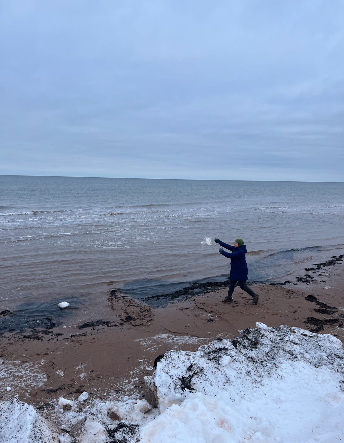 My wife, in hear winter coat and woolens, throws a big chunk of snow into the cold grey waters of Brackley Beach.