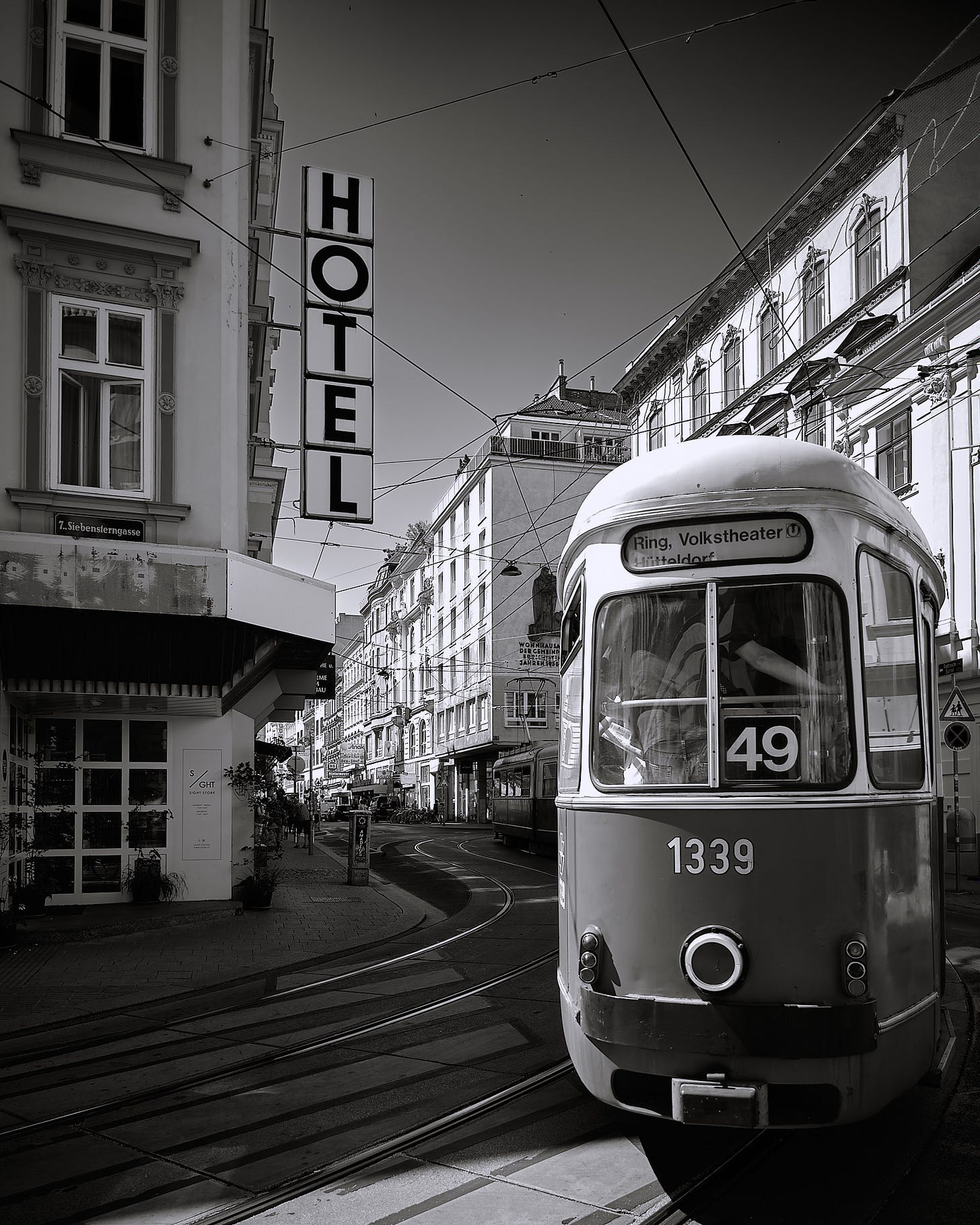 Black and white photograph of a Vienna cityscape featuring a tram marked '49' approaching a stop. Prominent 'HOTEL' sign is displayed vertically on a building to the left, while tram tracks curve through the cobbled streets and historical buildings line the backdrop.