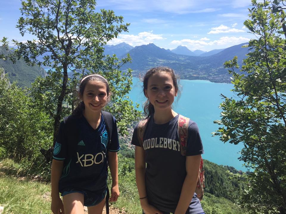 Two young girls smiling at the top of Mont Veyrier, overlooking the turquoise waters of Lake Annecy in France, with a backdrop of mountains and a clear blue sky.