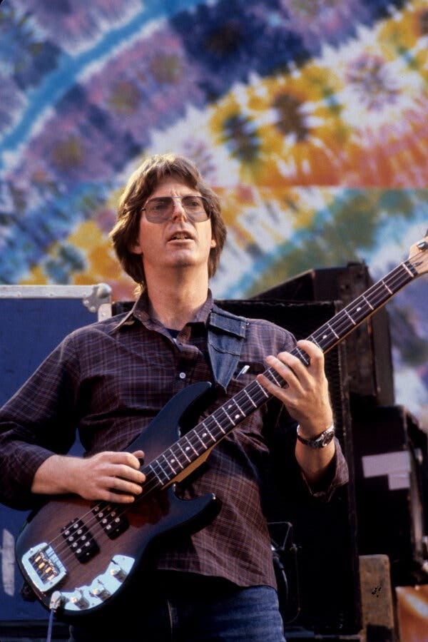 A portrait of Phil Lesh holding his bass guitar and standing against a colorful tie-dye background.