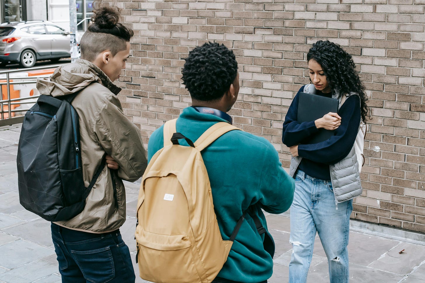 Stock photo, showing three teens having a conversation, and one of them averting their gaze to the floor as the others approach. 