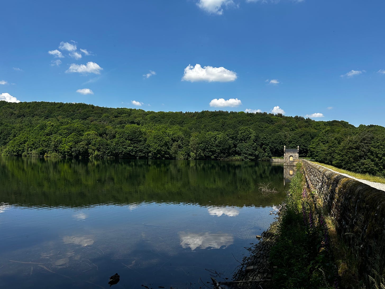 A photo of Linacre Reservoir, Cutthorpe, Derbyshire. The dam wall is on the right and the clouds are reflecting on the water. The lush green of the woods on the opposite shore look good in the sunshine. Image: Roland's Travels