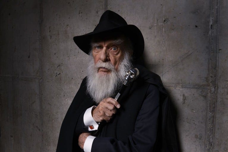 James Randi poses for a portrait against a concrete wall wearing a black brimmed hat and holding a cane with a skull topper