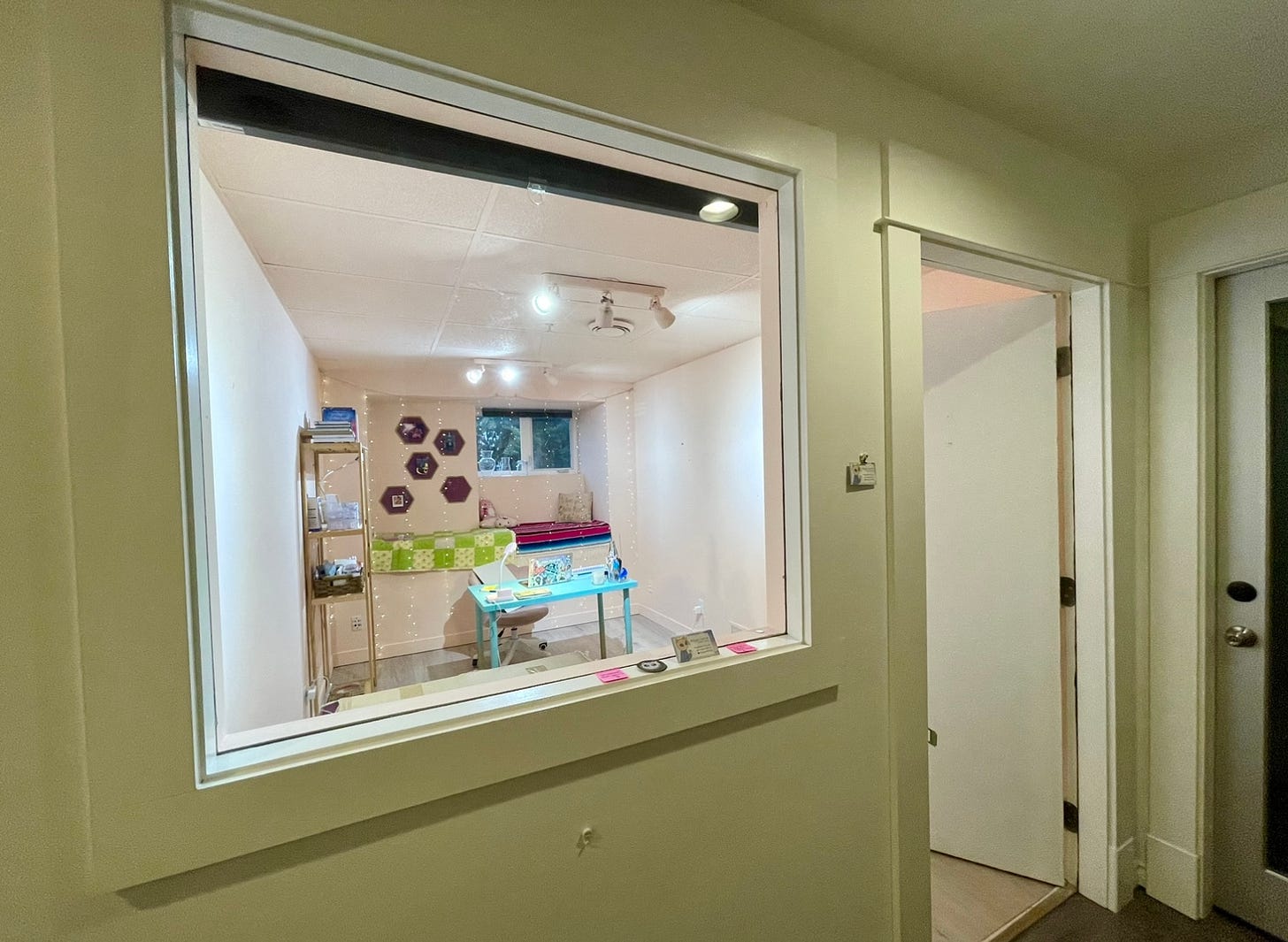 Image shows an open door and the view through an interior window that looks into a studio with pink walls, shelves, a teal desk and a nook beneath a window.