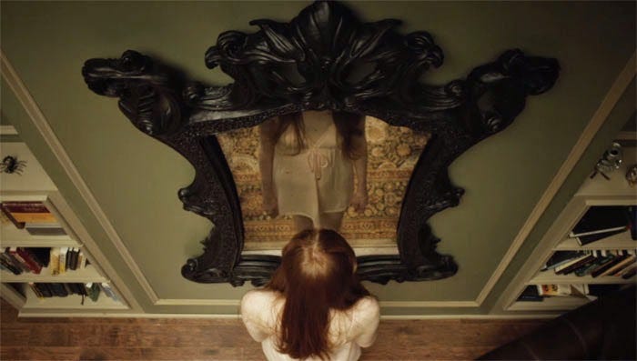 Scena del film oculus in cui una donna dai capelli rossi con indosso una camicia da notte rosa pallido sta davanti allo specchio antico. La scena è rispresa dall'alto