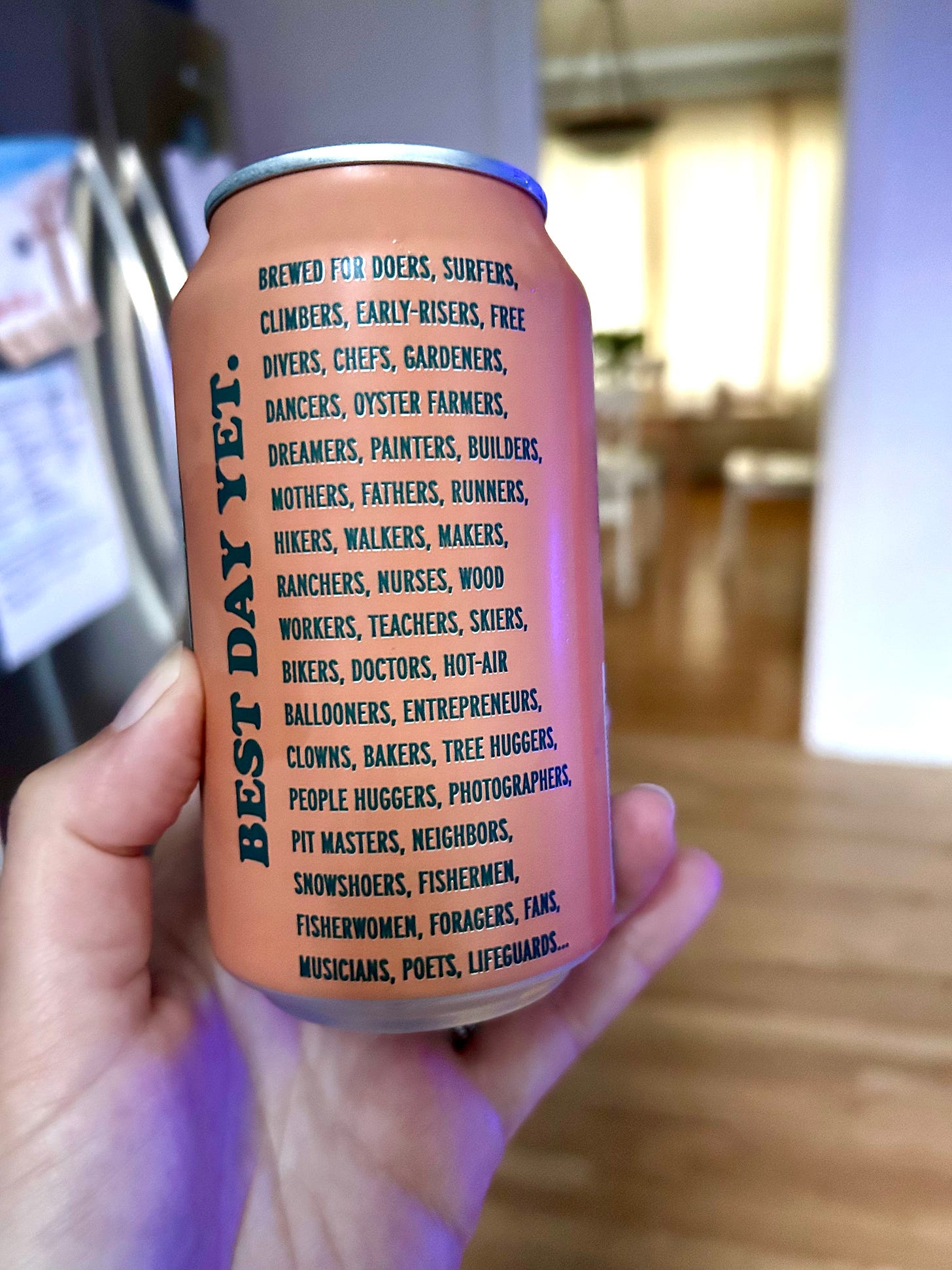 Image shows a hand holding a can of Best Day Brewing’s Kölsch-style beer with the back of the can showing. The can says “Best day yet,” followed by “Brewed for doers, surfers, climbers, early-risers, free divers, chefs, gardeners, dancers, oyster farmers, dreamers, painters, builders, mothers, fathers, runners, hikers, walkers, makers, ranchers, nurses, wood workers, teachers, skiers, bikers, doctors, hot-air ballooners, entrepreneurs, clowns, bakers, tree huggers, people huggers, photographers, pit masters, neighbors, snowshoers, fishermen, fisherwomen, foragers, fans, musicians, poets, lifeguards…”