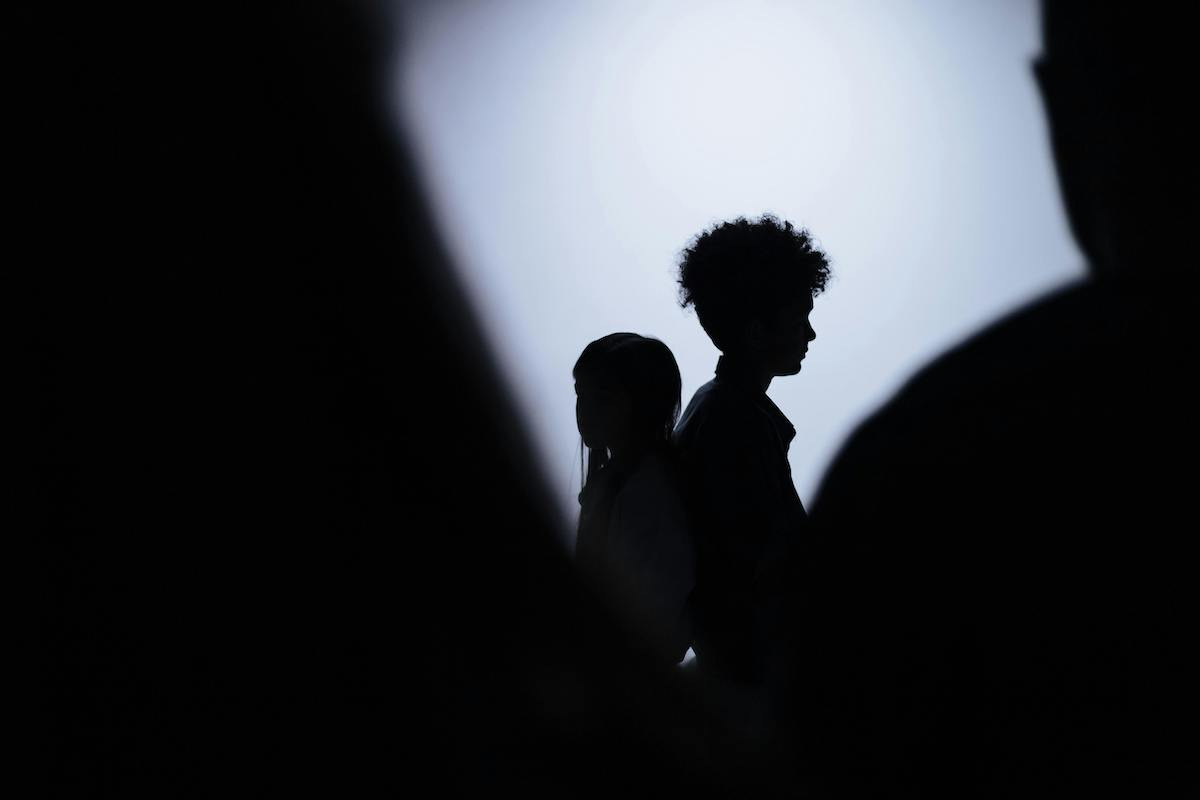 Photo of a silhouetted girl and boy standing back to back. In the foreground are silhouettes of two larger figures that seem to loom over the children – the one on the right appears to be an adult man.