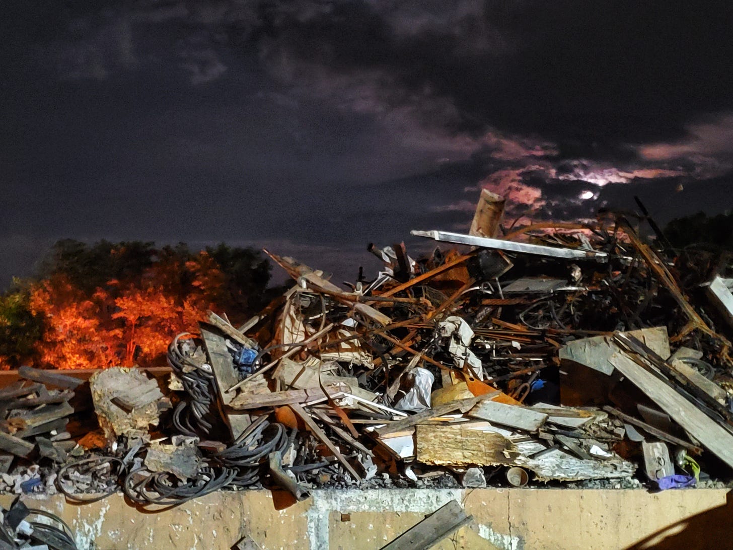 A pile of twisted and burned rubble rises in the foreground. The orange reflection of a fire lights up a cluster of trees in the background. Overhead, the night sky is lit by a nearly full moon.