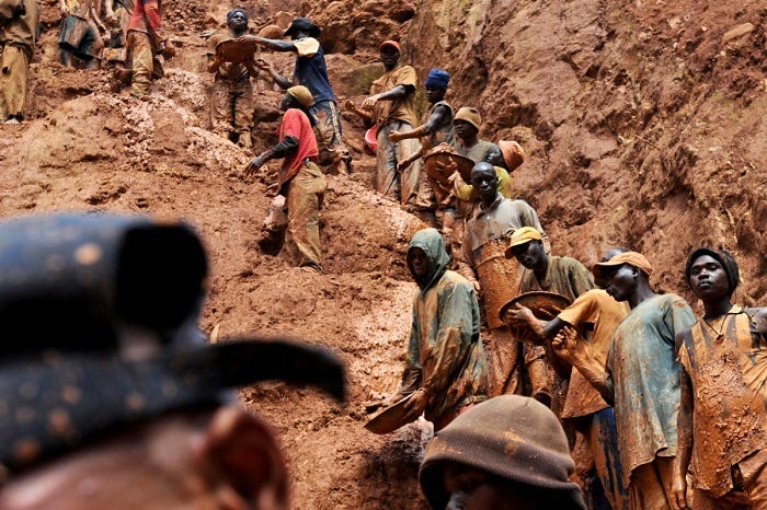 nineteen people within frames stand on a golden, muddy cliff as they work a gold mine in northeastern Congo. Many of them are covered in golden mud. the mountain “path” curves upward and out of frame, indicating there are more people the camera did not capture.