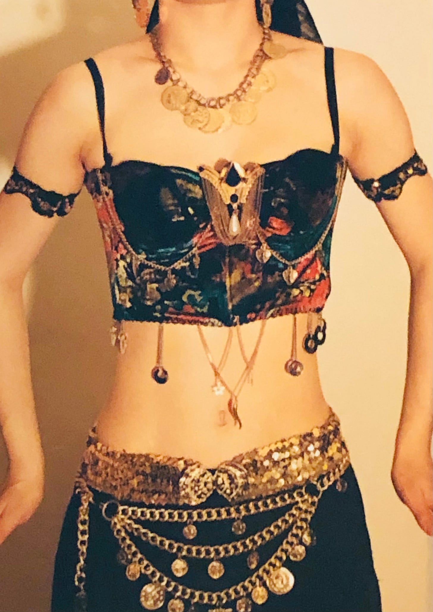 A black, red, green and gold bustier with chain adornments and golden dangles, a fancy gold brooch, gold sequin belt over a belt of gold chain drapes laden with coins.