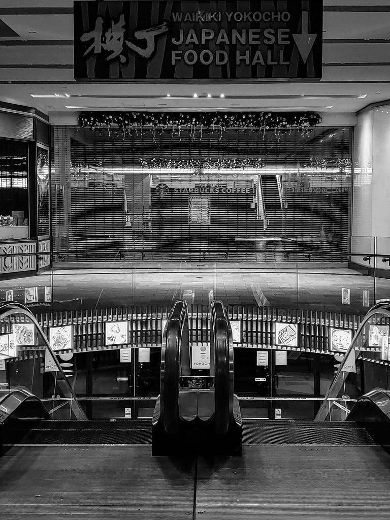 Monochrome photo showing escalators to a Japanese food court in Waikiki. A sign at the top of the frame reads "Japanese Food Hall." Immediately below the sign is a roll-down metal gate at the entrance to a shopping mall. Two stores are shown in partial profile to the right and left of the gate. In front of the gate is a cobblestone entry. In front of the escalators is a paving stone sidewalk.