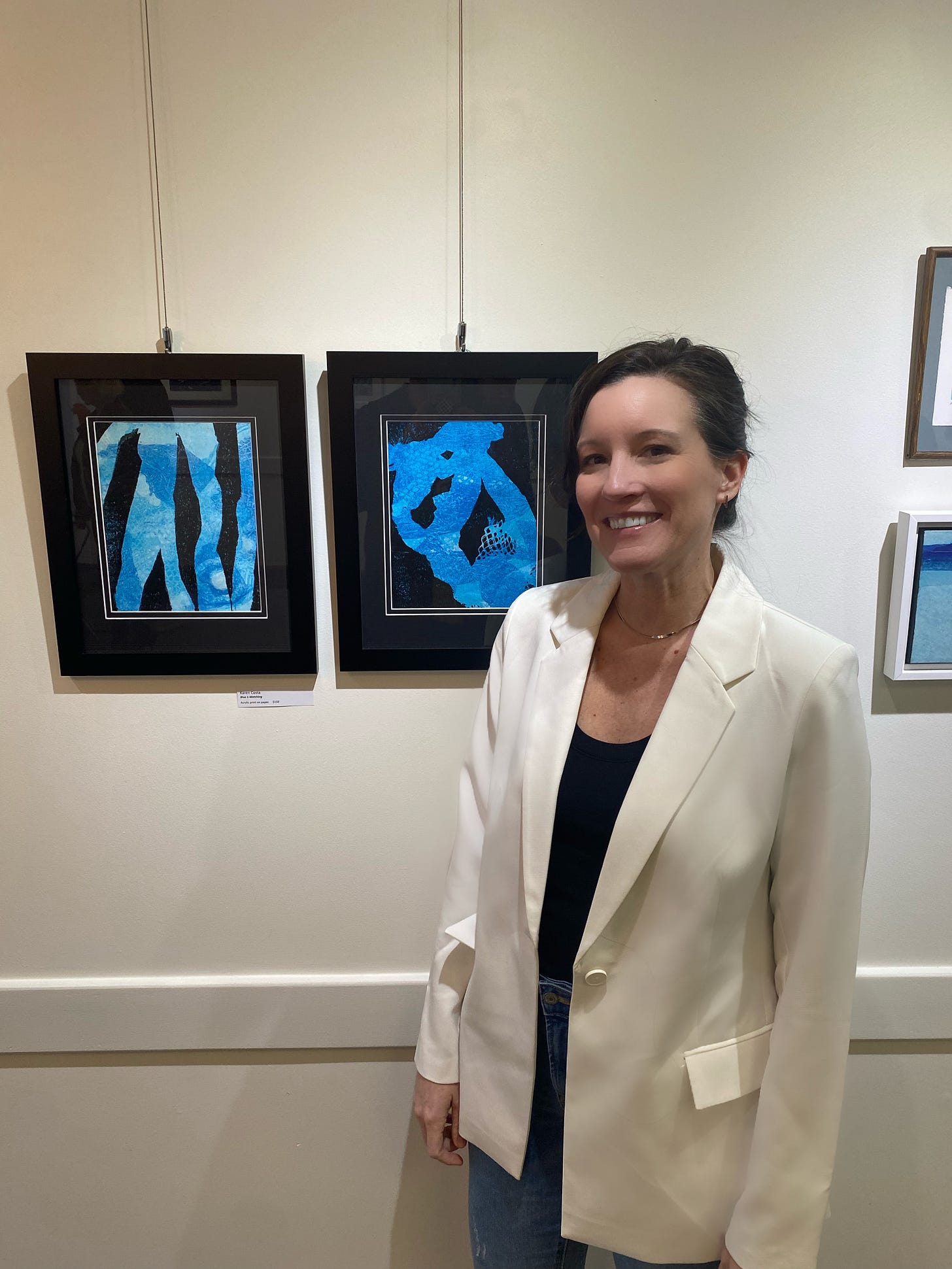 Two blue and black prints hang on an art gallery wall. Karen Costa, smiling, stands next to them wearing a white blazer.