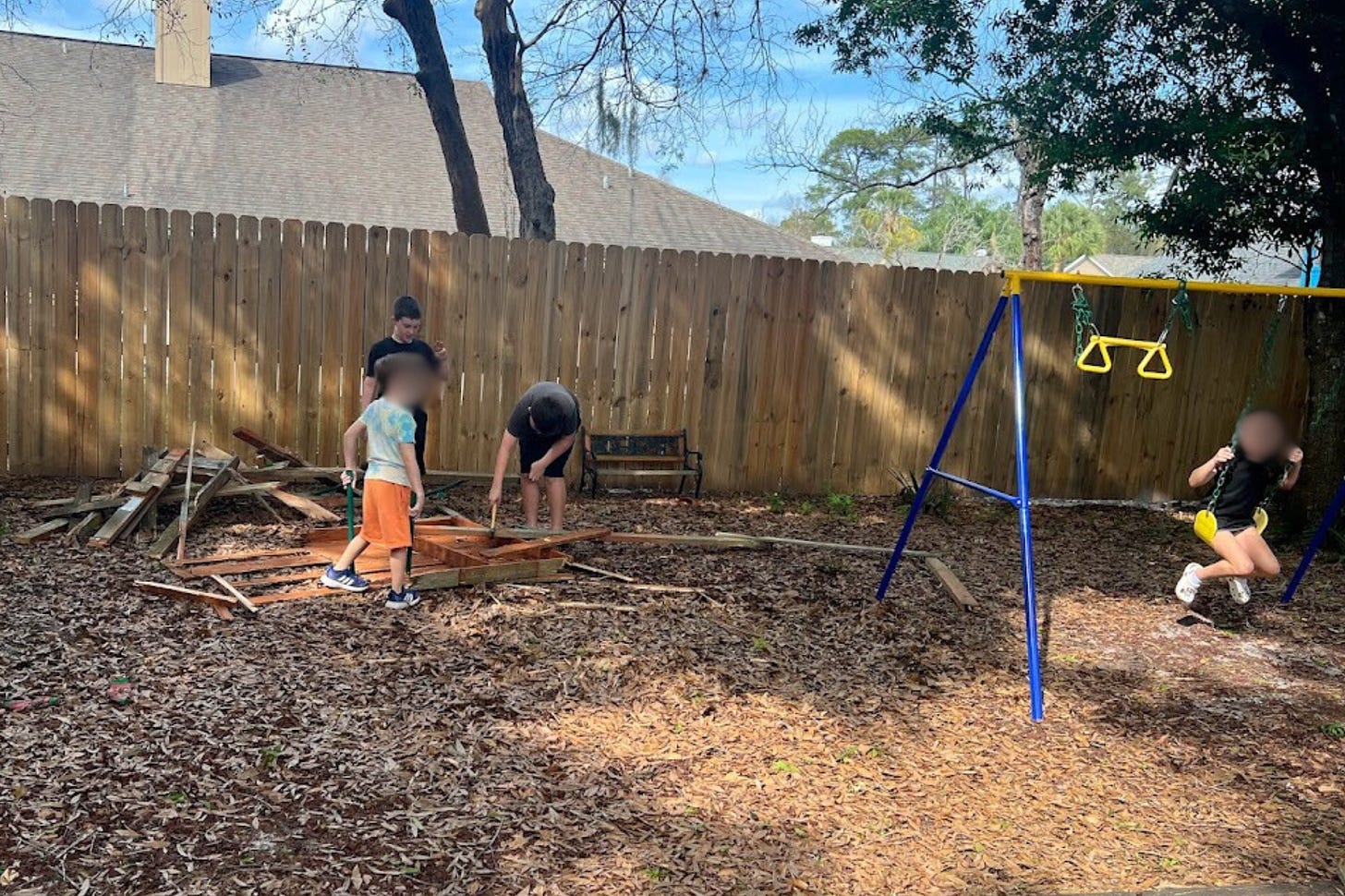 A partially dismantled wooden playground structure in a backyard, with children engaged in what appears to be demolition work. A child is seen swinging nearby, suggesting a juxtaposition of play and work in the open, sunlit space bordered by a wooden fence and trees.