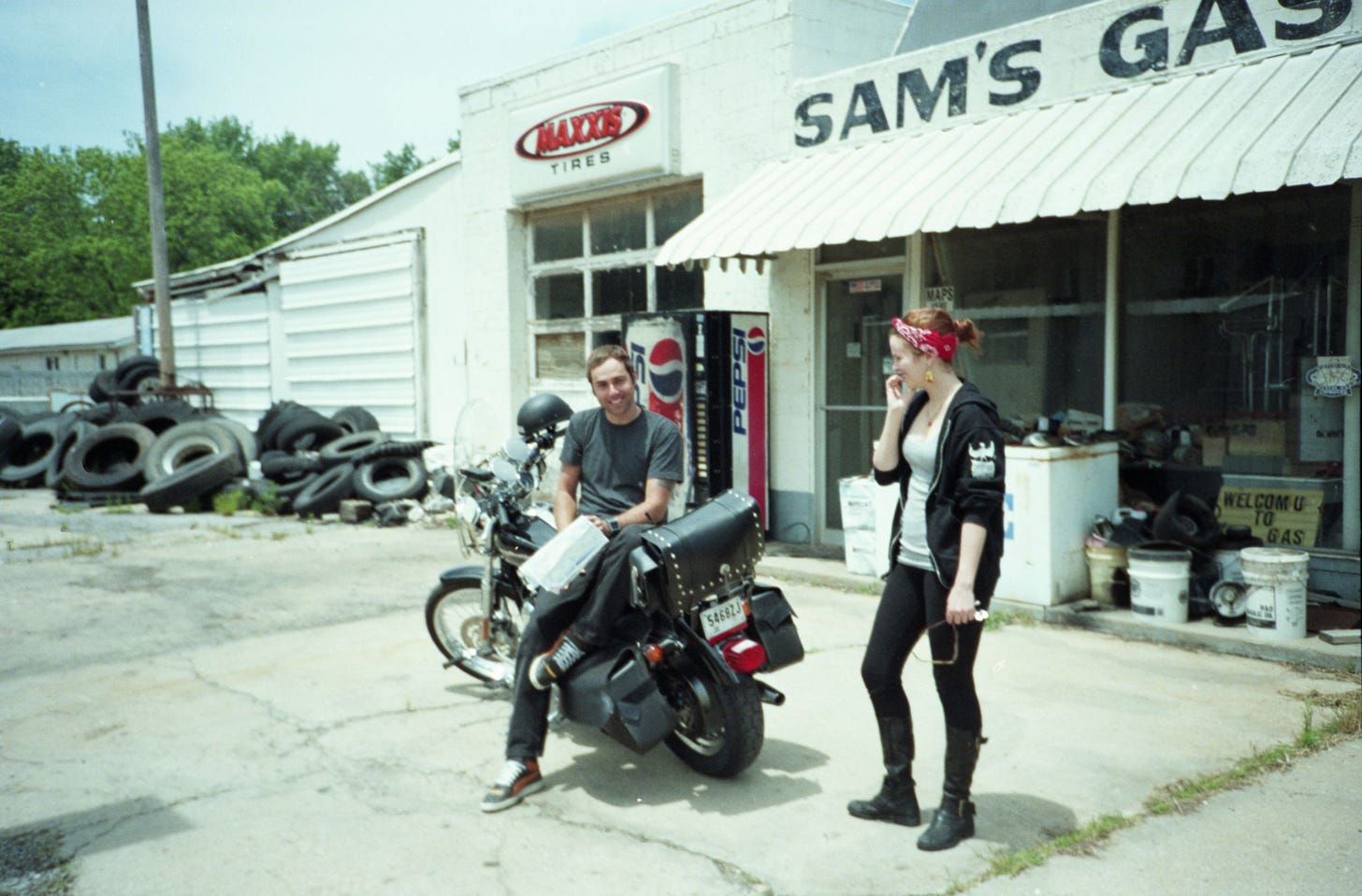 Derrick sits on the parked bike. He looks into camera, smiling slightly. Amber stands near him, looking at him. They are in front of a rundown looking storefront called "Sam's Garage." There is a pile of tires in the background. 