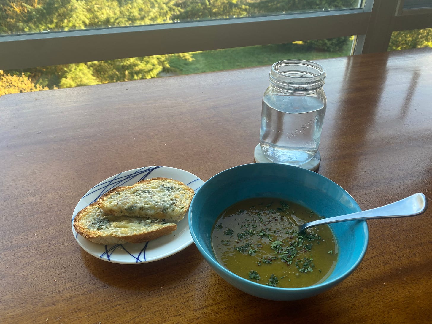 A blue bowl of light brown lentil & squash soup, topped with chili flakes and chopped cilantro, a spoon sticking out of the bowl. Next to it is a small white plate with cheesy toast, browned at the edges and with the cilantro visible beneath the white cheddar. A mason jar of water sits on a coaster, and the light in the trees is just visible out the window at the top of the frame.