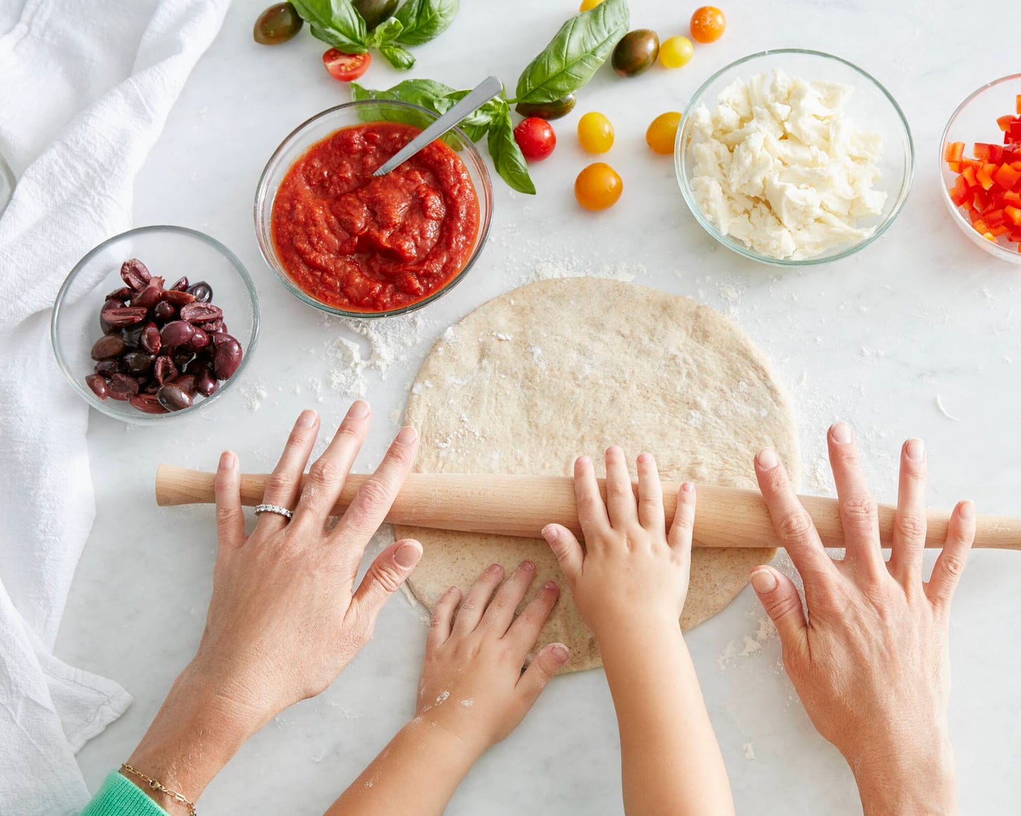 Adult hands and kid hands rolling out homemade pizza dough, with pizza sauce and cheese in side bowls