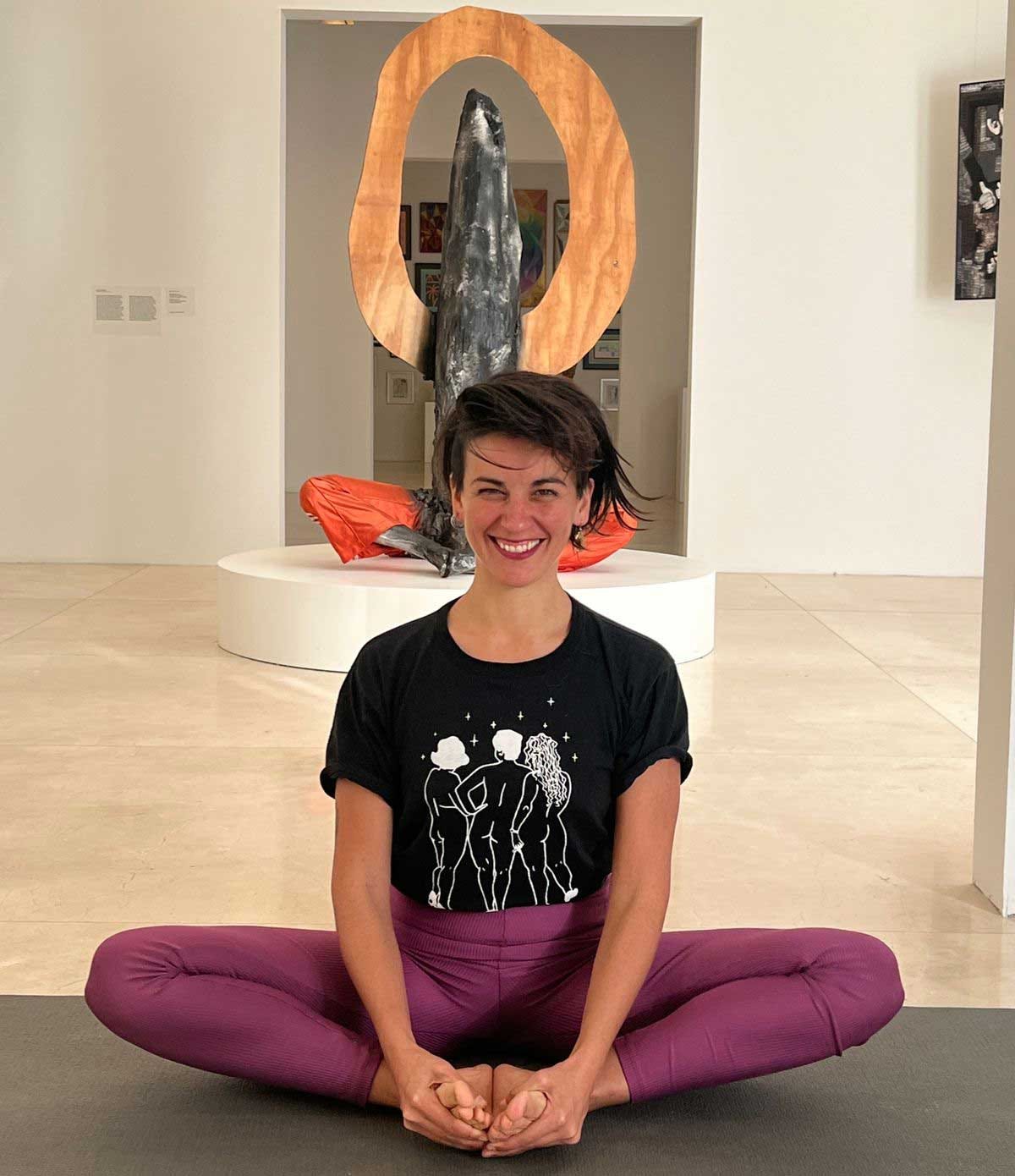 Photograph featuring an adult woman (Carolina) at the center of the image. She is seated in a simple yoga pose on a mat inside a contemporary art museum gallery. Behind her, there is a sculpture made of wood and metal that mirrors the posture she is holding on the floor. Photograph featuring an adult woman (Carolina) at the center of the image. She is seated in a simple yoga pose on a mat inside a contemporary art museum gallery. Behind her, there is a sculpture made of wood and metal that mirrors the posture she is holding on the floor.