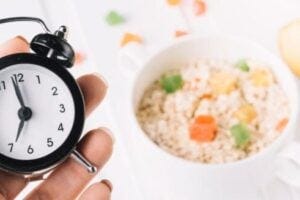 A hand holding a small clock in front of a meal