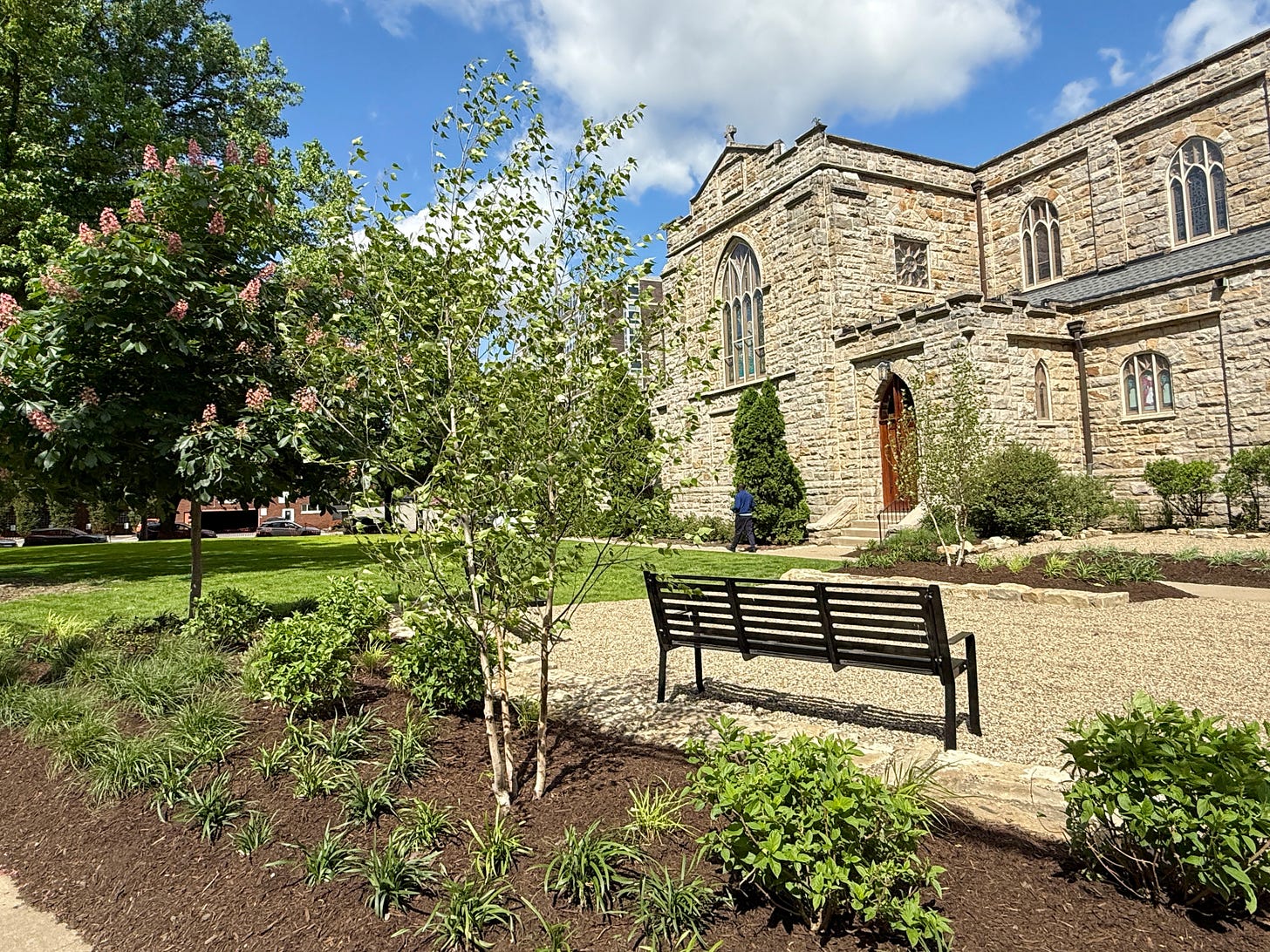 Looking towards the nave door from sidewalk over green trees and grasses with mulch, and black metal benches on pea gravel.