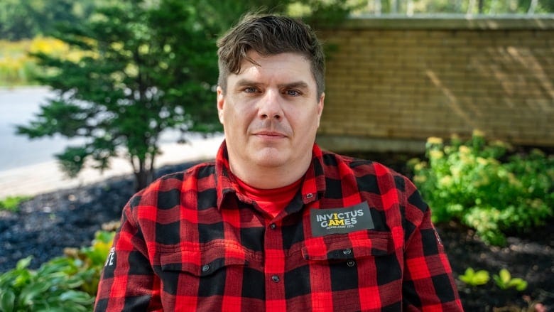 A man in a black and red flannel occupies the frame, the Invictus Games badge over his left breast, as he stares into the camera. A man in a black and red flannel occupies the frame, the Invictus Games badge over his left breast, as he stares into the camera.