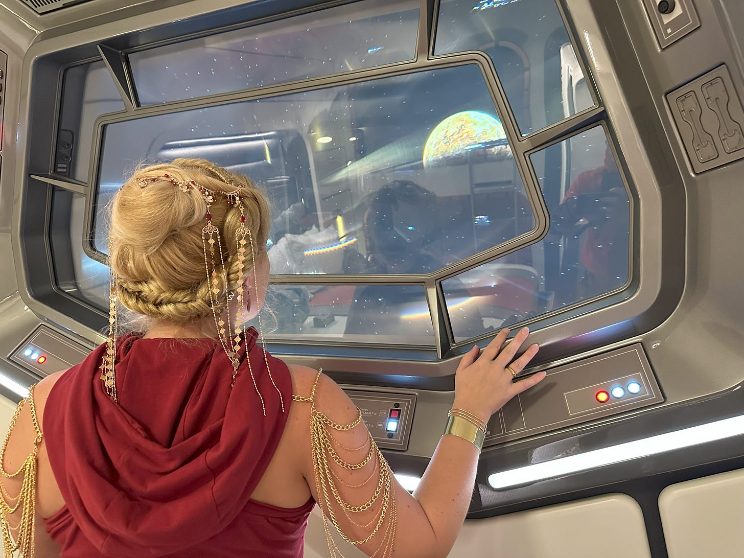 Cass standing with her back to the camera, looking out the viewscreen of the Halcyon passenger room, which shows the planet Chandrila. She is wearing a red hooded dress; her hair is elaborately styled with golden jewelry, and she wears gold shoulder jewelry and bracelets.