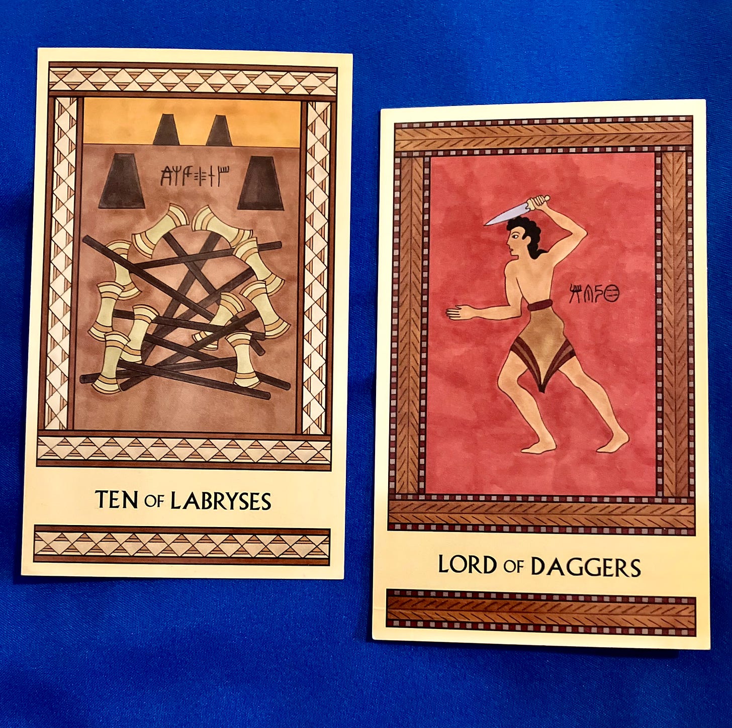 Two Minoan Tarot cards on a royal blue background. The Ten of Labryses is in shades of gold and tan. It shows ten labryses in a pile on the floor with four empty labrys stands in the background. The Lord of Daggers is in shades of deep red and brown. It shows a Minoan man, standing, facing left, brandishing a dagger over his head.