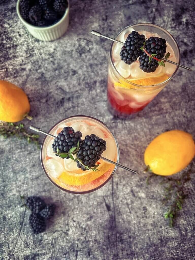 Top down view of two purple and gold mocktails in tall skinny glasses, garnished with a lemon wheel and skewered blackberries