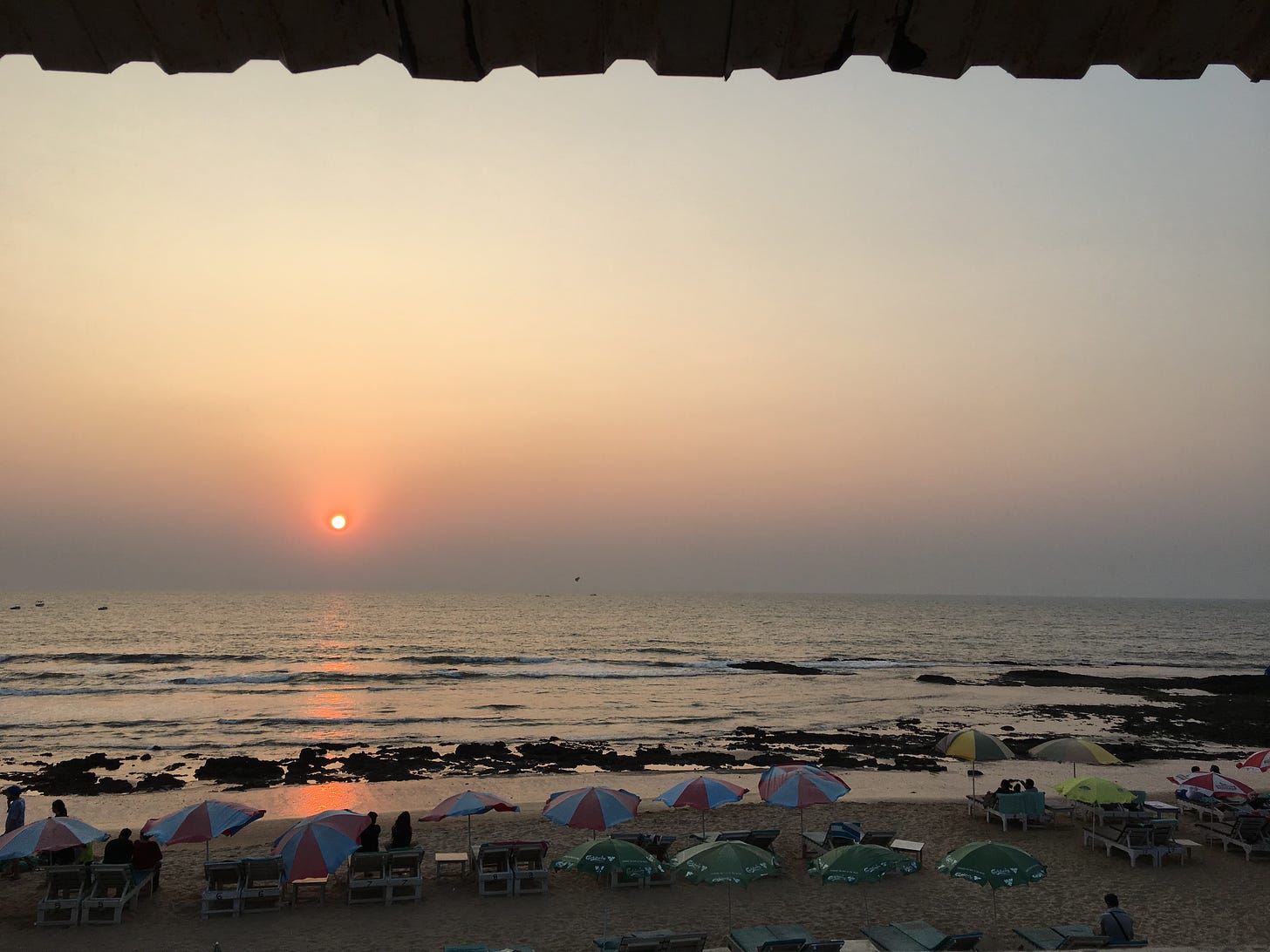 The sun sets over a beach. On the shores, crowds sit on beach chairs and under umbrellas to admire.