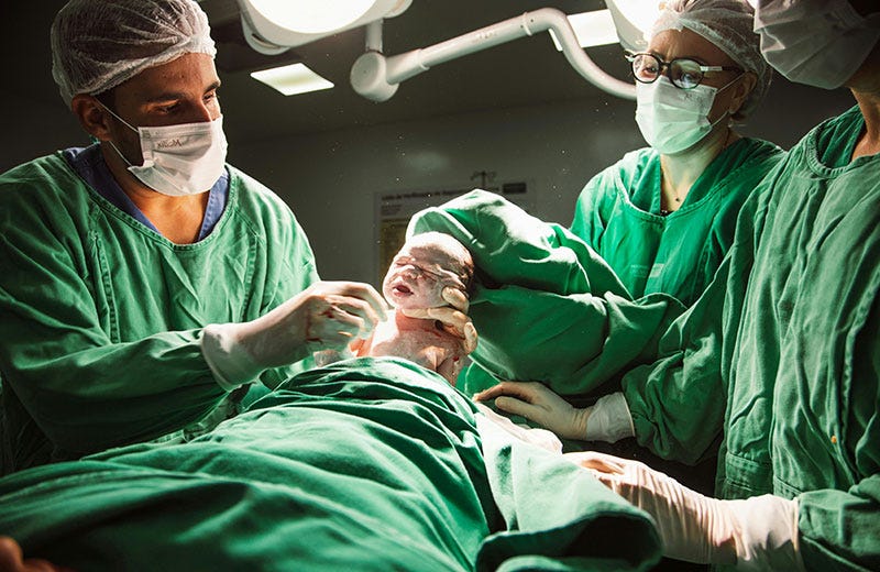 A photo of medical staff in teal scrubs lifting a newborn out of the uterus during a cesarean birth.