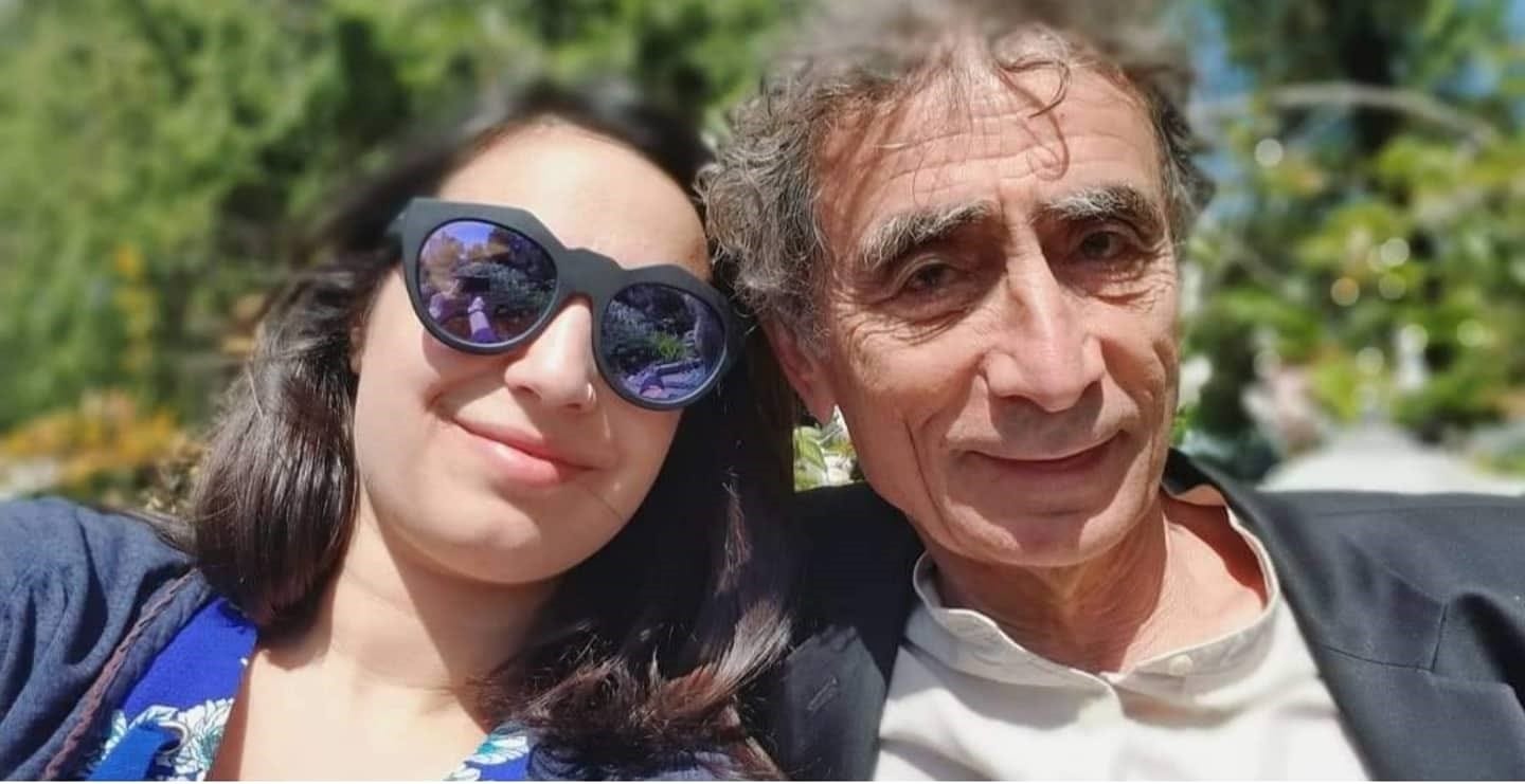 Hannah and Gabor Mate sit together in the sunshine against a backdrop of trees. Hannah is wearing dark sunglasses; their heads are inclined towards one another. Both are smiling.