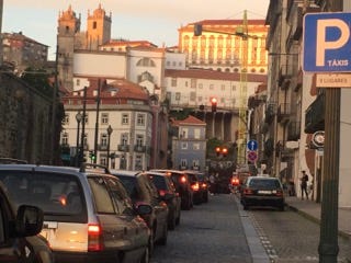 A sign indicates a taxi stand beside a car-filled, cobbled road with old buildings and a two-turreted building, perhaps a church, in the background, in the city of Porto, all gilded by the golden hour.