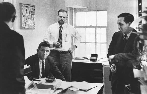 A black-and white photo of three men at a desk, two of them standing and the other (a young Norman Mailer) sitting. Mr. Fancher wears a shirt and tie but no jacket and has his left hand on his hip.