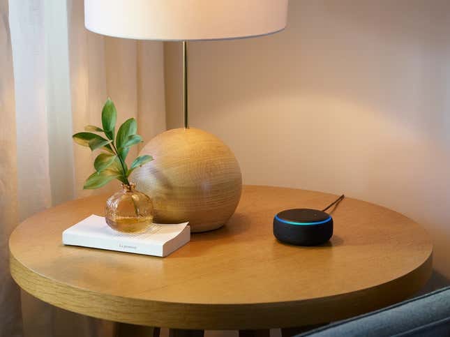 Echo Dot sitting on a light brown wood table beside a lamb and book and fake plant