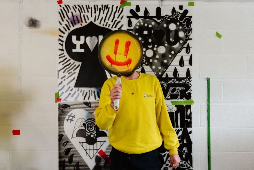 A photograph of a person wearing  a yellow sweatshirt, holding a yellow smiley face up in front of their own face.