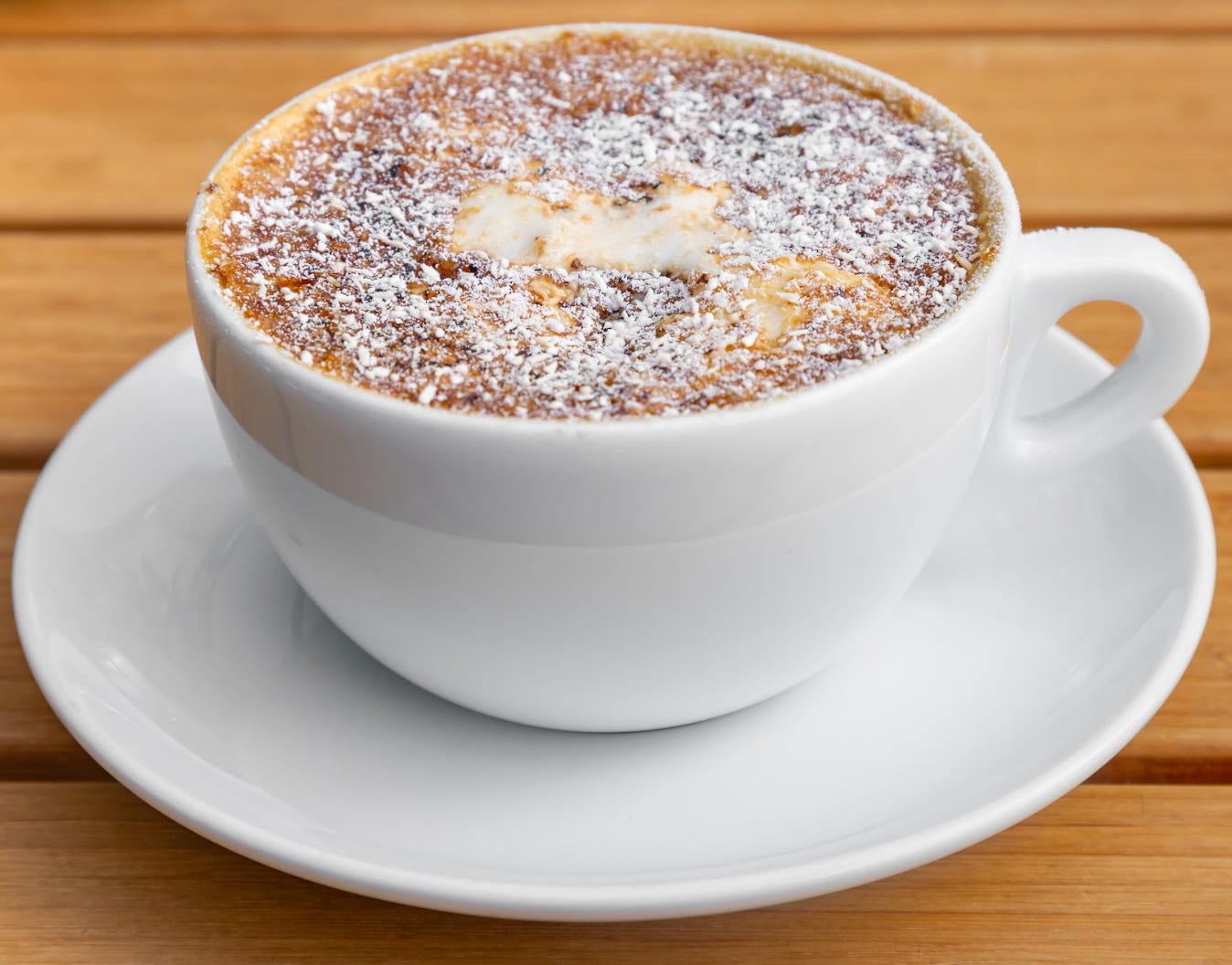 Close up from above of a creme brulee latte topped with powdered sugar in a white coffee mug on a white saucer on a light wood tabletop.