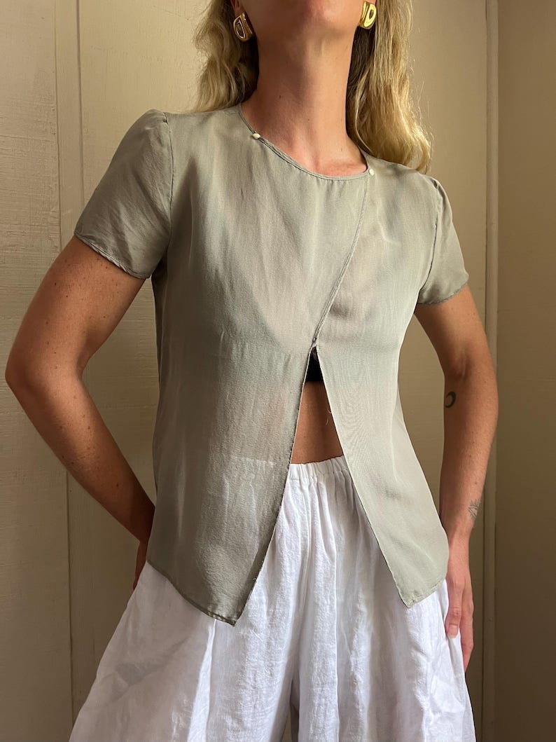 May include: A light green silk blouse with a short sleeve and a deep V-neckline. The blouse is semi-sheer and has a slightly cropped length.